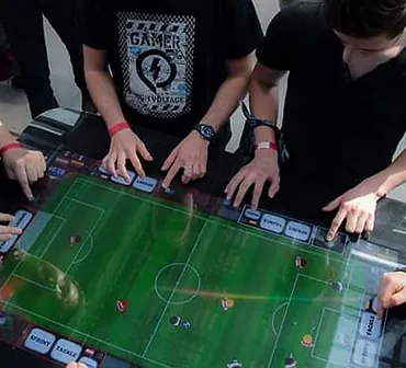 Stort digital spillebord med fodboldspil set fra oven. Rundt om ses hænder på bordet som spiller på touch skærmen